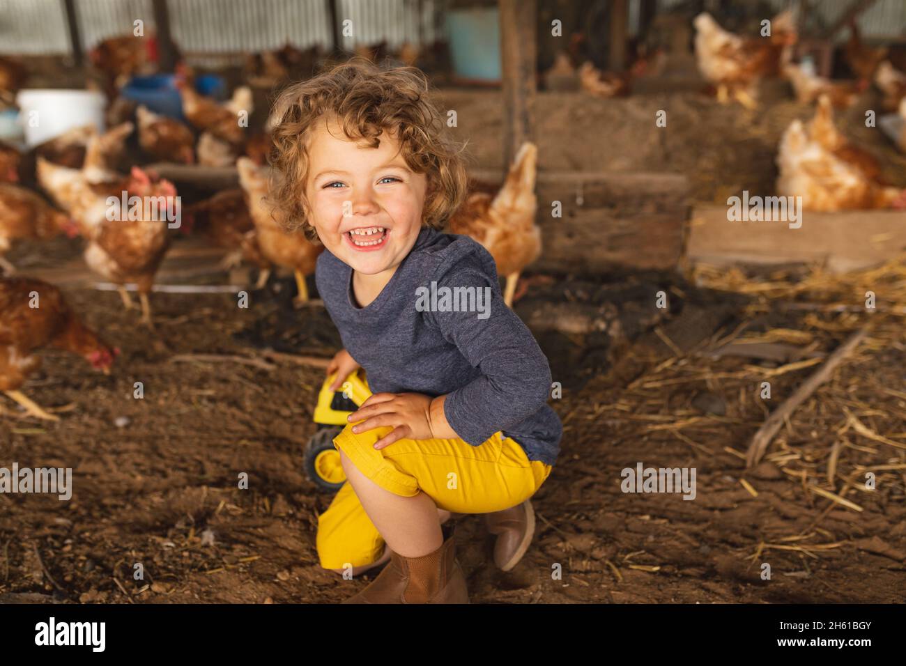 Porträt eines glücklichen niedlichen Jungen, der mit Hühnern im Hintergrund auf der Geflügelfarm kniet Stockfoto