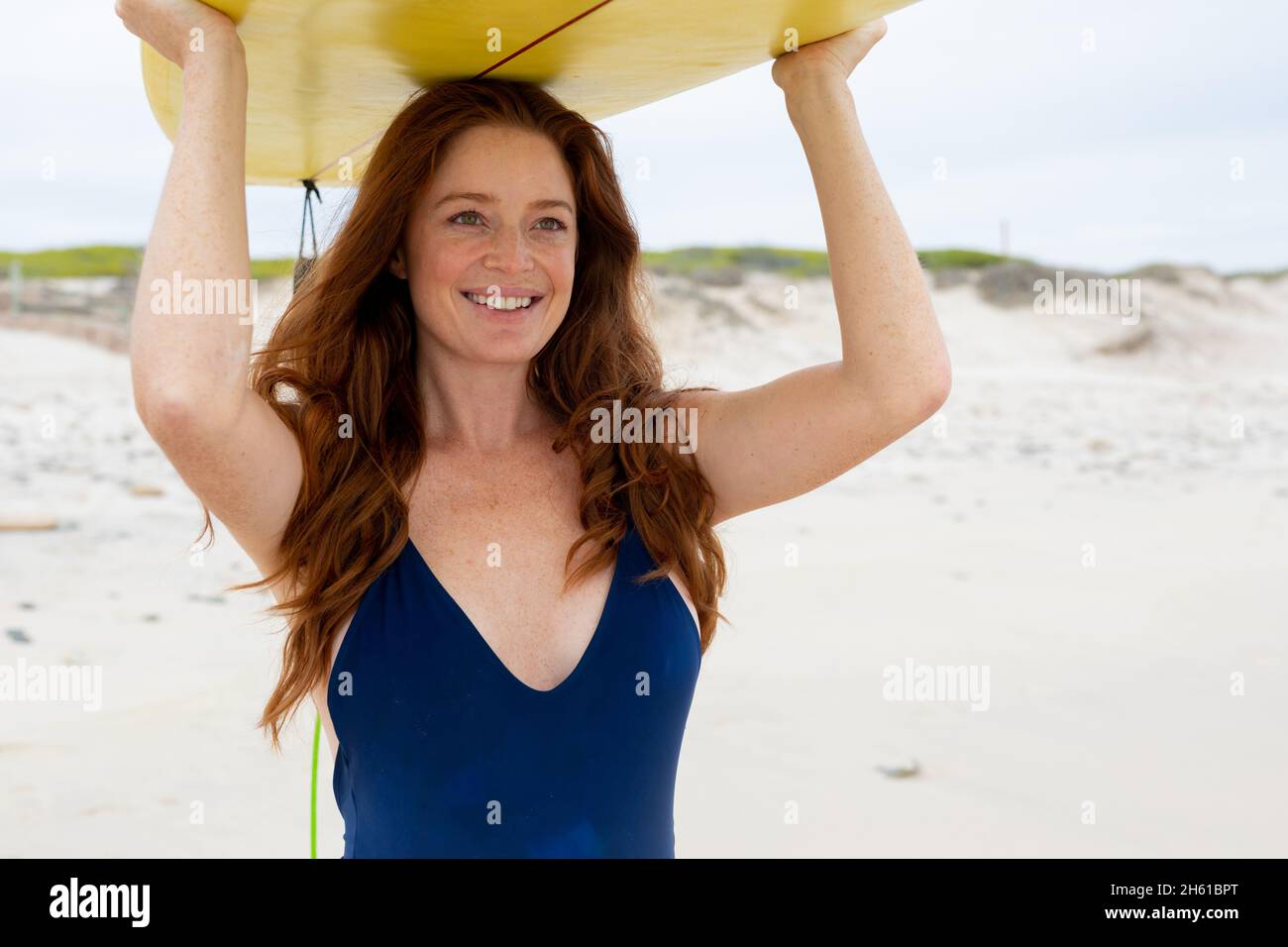 Lächelnde junge kaukasische Frau mit Surfbrett auf dem Kopf, während sie am Wochenende am Strand spazieren ging Stockfoto