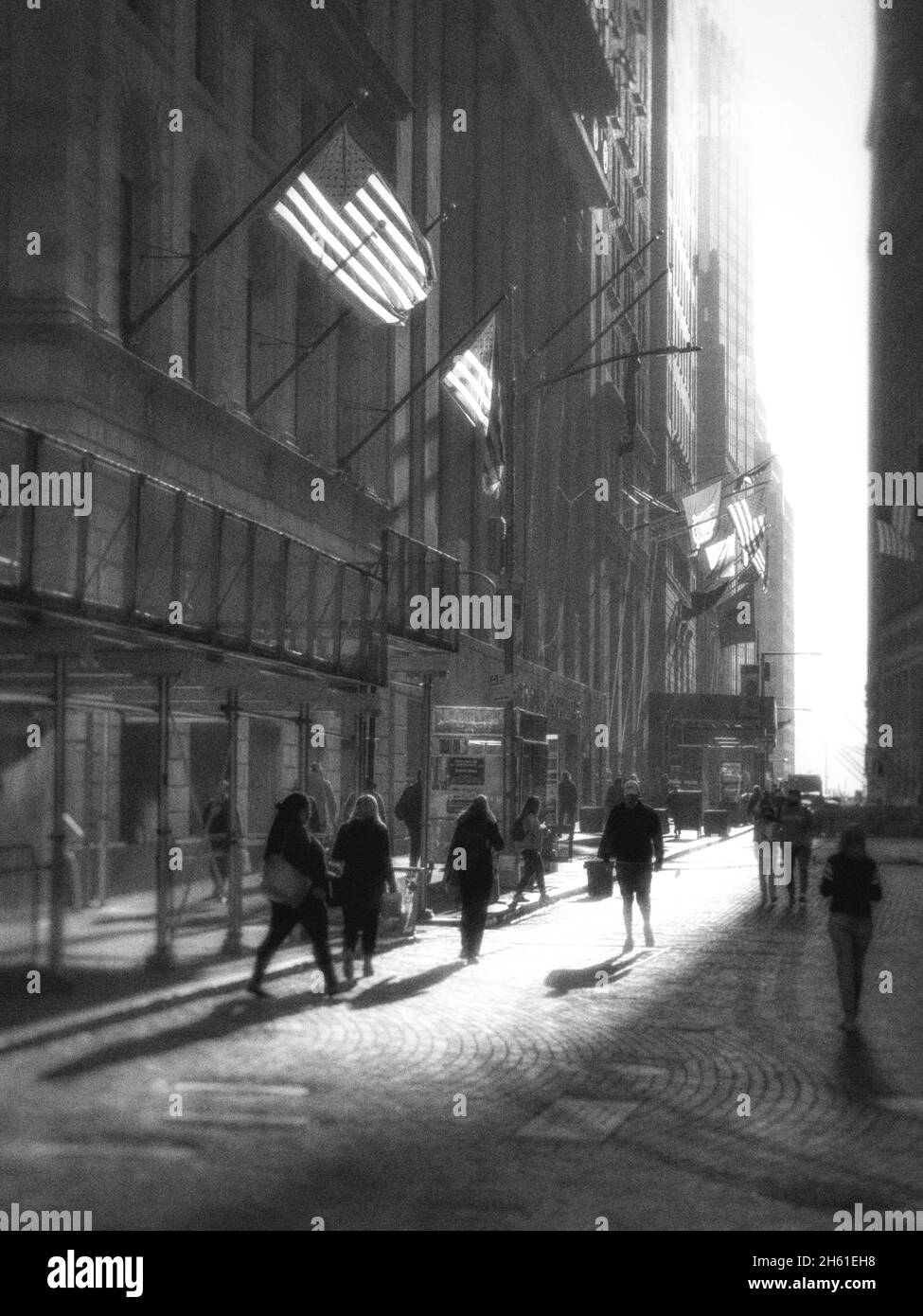 Silouette von Menschen in einer Kopfsteinpflasterstraße im unteren Manahattan in der Nähe der New Yorker Börse. Hinterleuchtete amerikanische Flaggen auf den Gebäuden Stockfoto