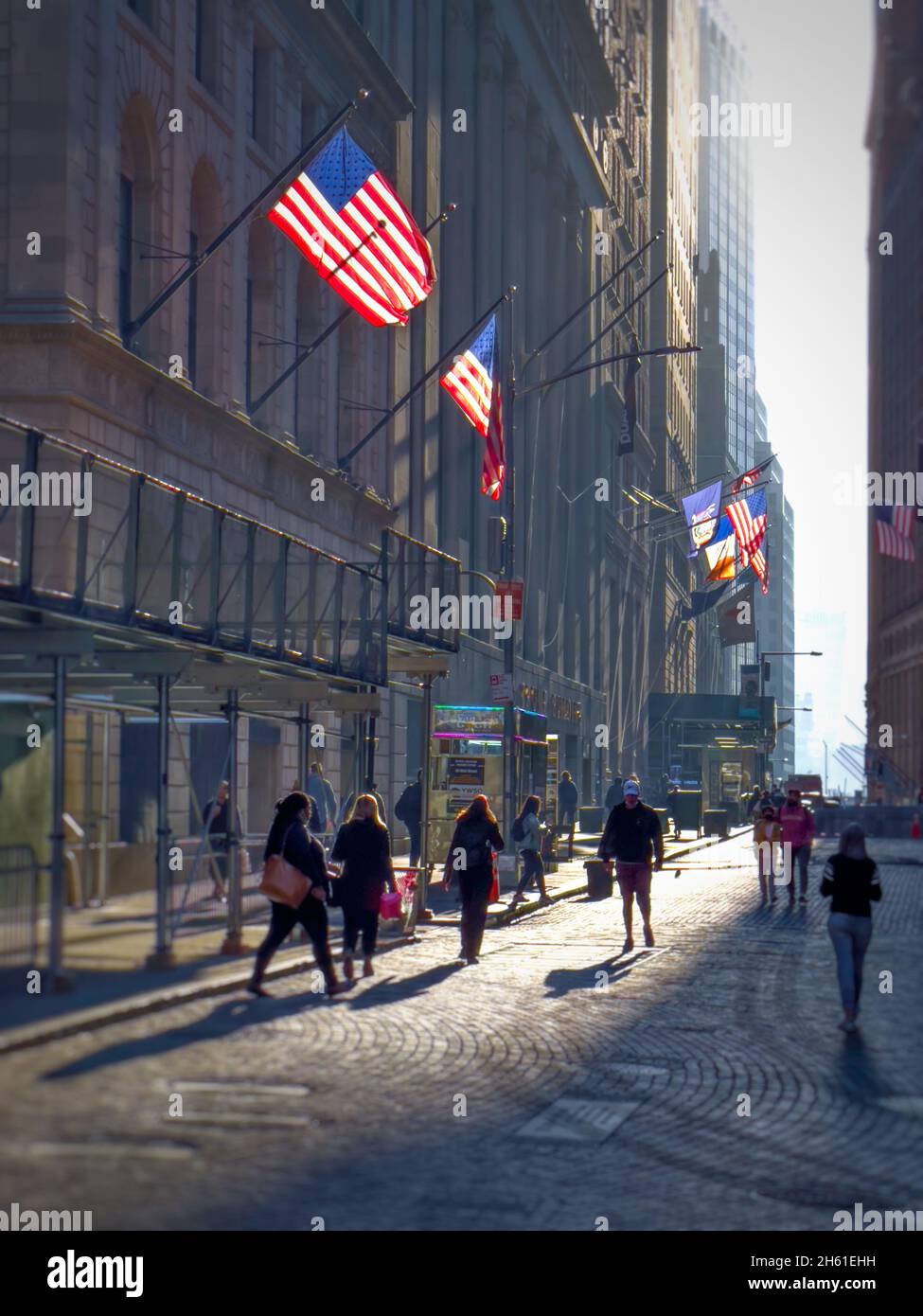 Silouette von Menschen in einer Kopfsteinpflasterstraße im unteren Manahattan in der Nähe der New Yorker Börse. Hinterleuchtete amerikanische Flaggen auf den Gebäuden Stockfoto