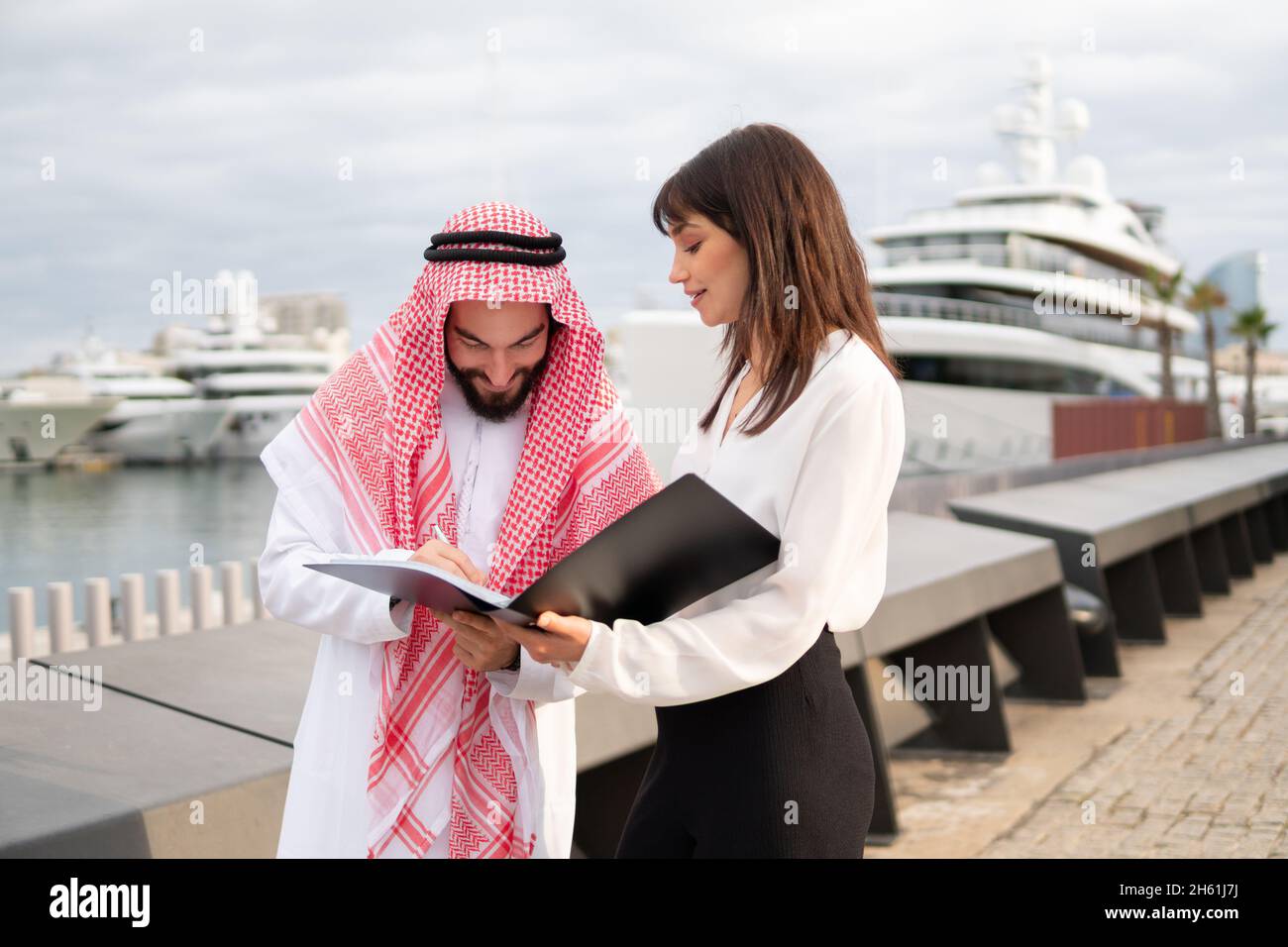 Ein lächelnder arabischer Geschäftsmann, der einen Vertrag mit einem Schreibergesang hält, während er mit einer jungen europäischen Geschäftsfrau im Hafen steht, ein arabischer Mann, der bei einem Treffen mit einem Geschäftspartner im Freien die Unterschrift auf die Dokumente legt Stockfoto