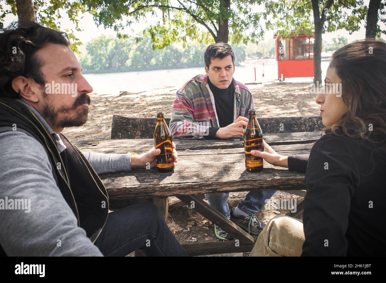 Eine Gruppe von drei unglücklichen jungen Freunden, weiblich und männlich, sitzen zusammen und trinkt Bier aus der Flasche auf einer Holzbank mit Tisch im Waldpark im Freien. Rea Stockfoto