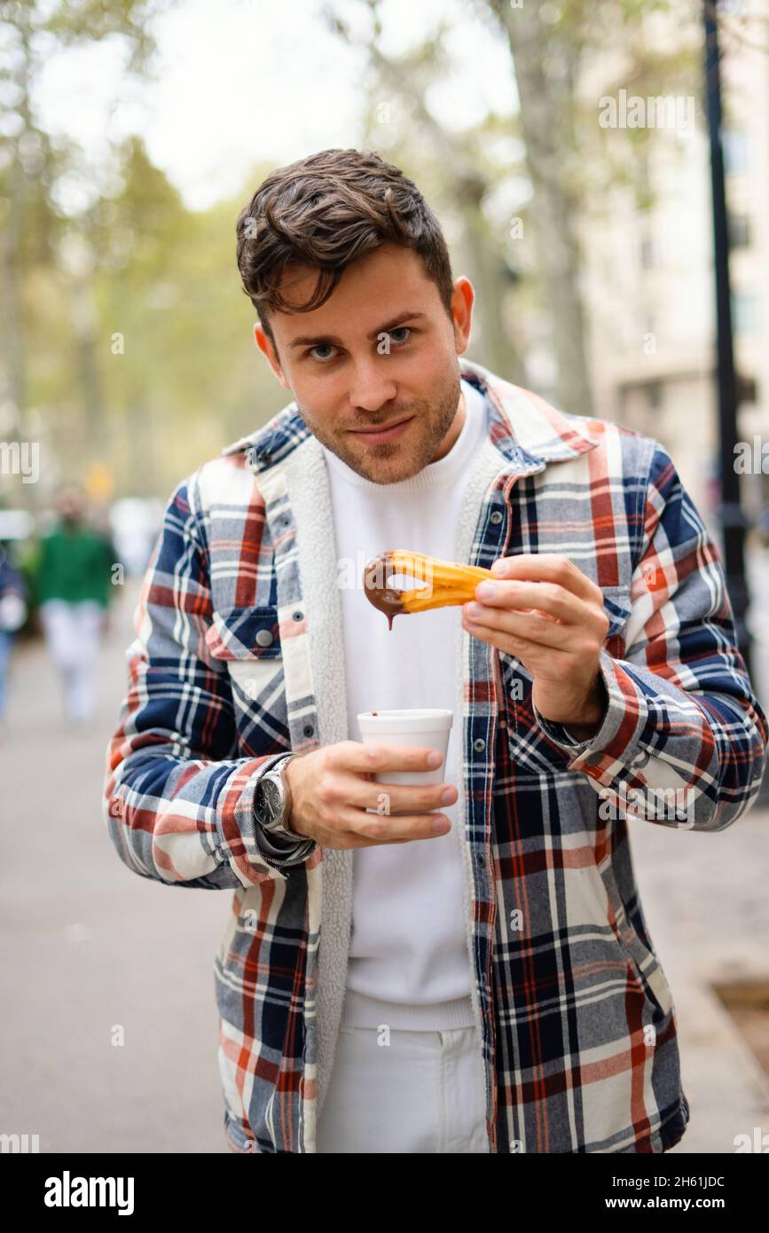 Lächelnder Mann, der süße Churros isst, während er mit einer Tasse heißer Schokolade in der Stadt steht und die Kamera anschaut Stockfoto