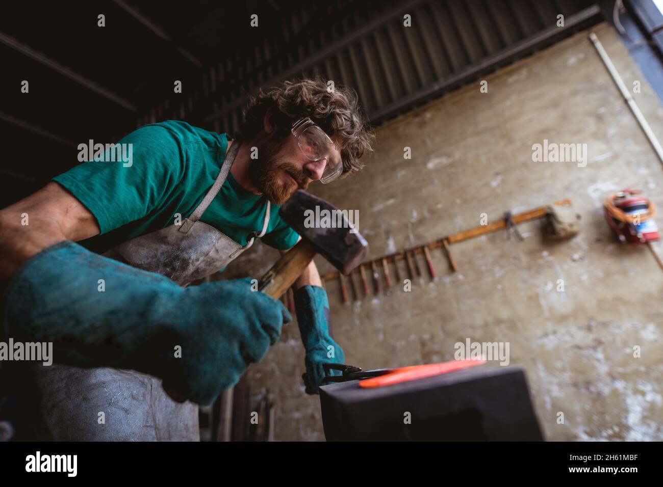 Bärtiger kaukasischer Schmied in Schutzkleidung Schmieden mit Hammer auf Amboss in der Industrie Stockfoto