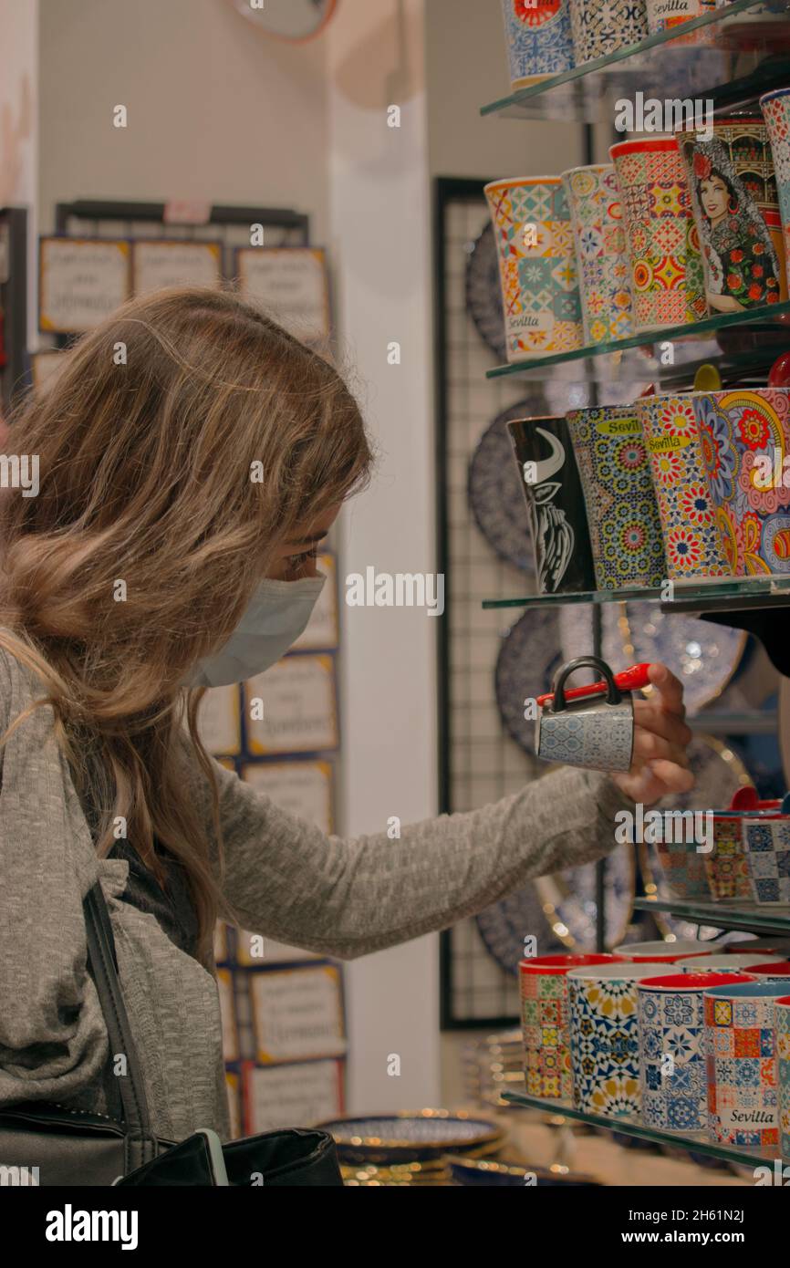 Kaukasisches Mädchen mit Gesichtsmaske auf Looks im Souvenirladen, um ihren Lieben ein Souvenir ihrer Reise nach Sevilla zu geben. Stockfoto