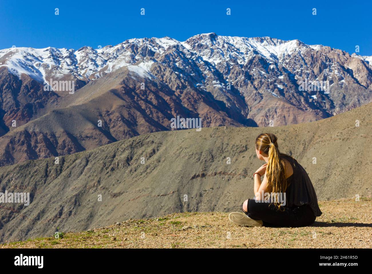 Junges blondes Mädchen, das auf einem kargen Boden sitzt und die schneebedeckten Berge der Anden unter blauem Himmel im Elqui-Tal, Chile, anschaut Stockfoto