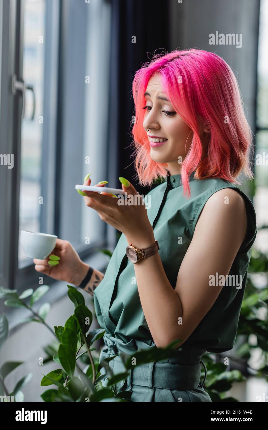 Fröhliche Geschäftsfrau mit rosafarbenem Haar und Piercing, die eine Tasse Kaffee hält, während sie die Sprachnachricht auf dem Smartphone aufzeichnet Stockfoto