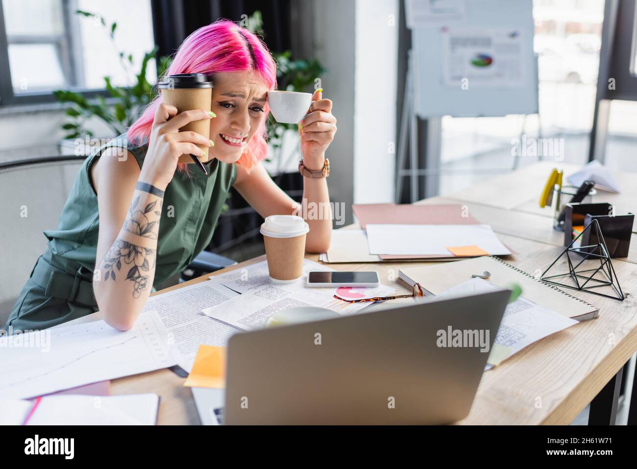 Besorgte Geschäftsfrau mit rosafarbenem Haar, die Kaffee hält und auf den Laptop schaut Stockfoto