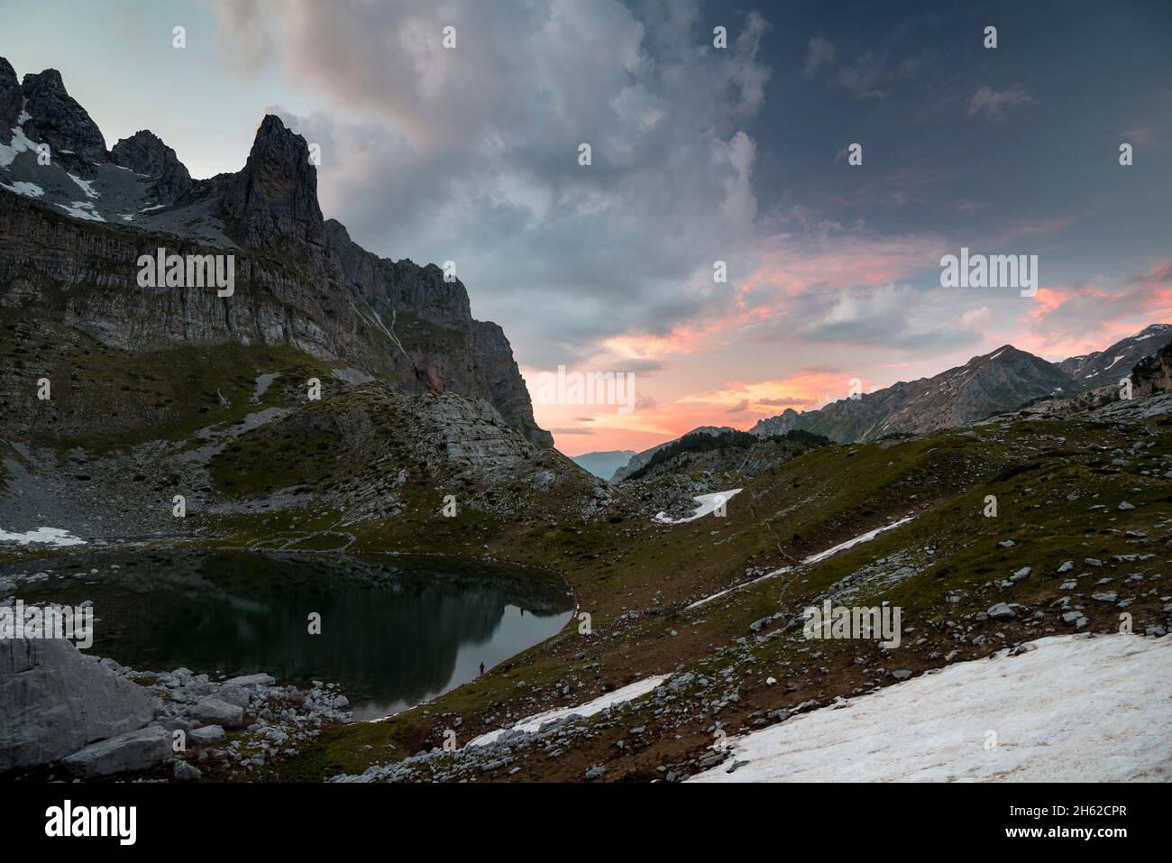 Eine einzelne, kaum sichtbare Person steht am Rande eines Bergsees und blickt auf den Sonnenuntergang in der rauen Landschaft, montenegro, albanien, prokletije Nationalpark Stockfoto
