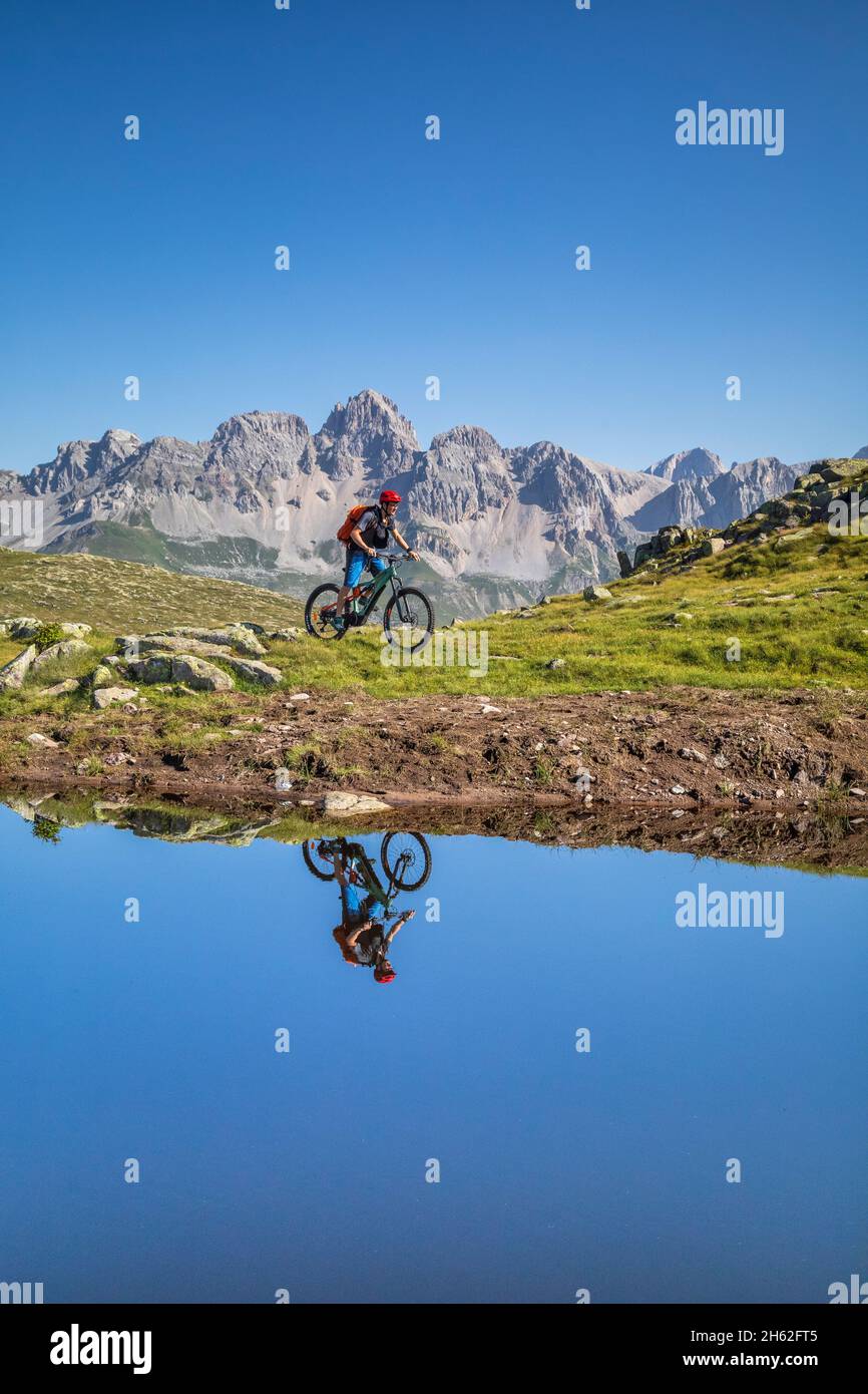 Mann, 22 Jahre alt, Radfahrer mit E-Bike in der Nähe eines alpinen Sees, Radfahrer im Wasser eines Bergsees reflektiert, Berge im Hintergrund. Laresei, falcade, belluno, veneto, italien Stockfoto