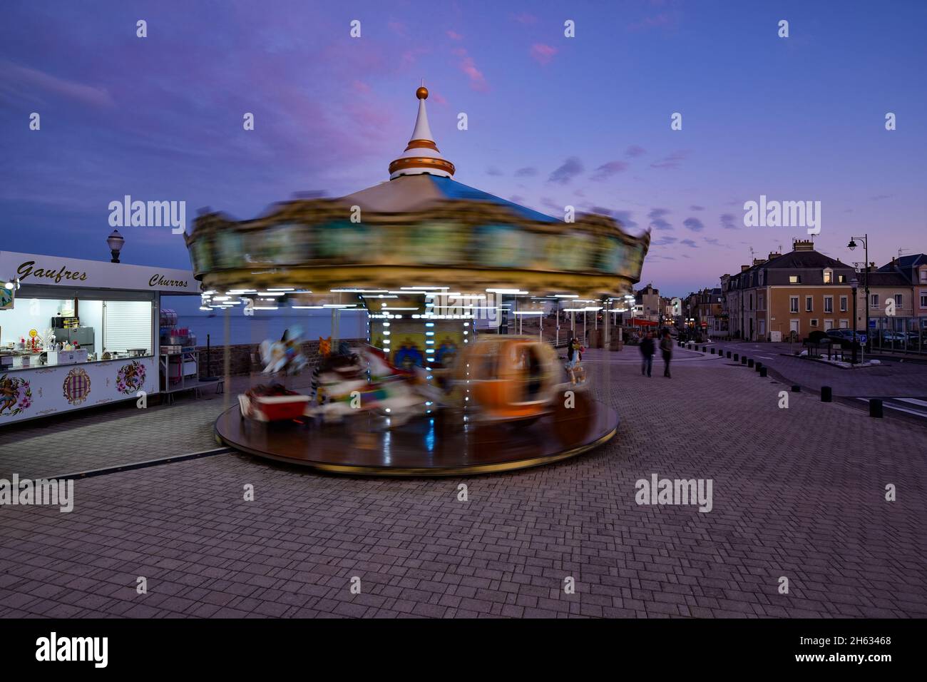 Strandpromenade in Saint-Aubin-sur-Mer zur blauen Stunde mit einem schönen Kinderkarussell in Bewegung und einem Süßigkeitengeschäft im Vordergrund. Stockfoto