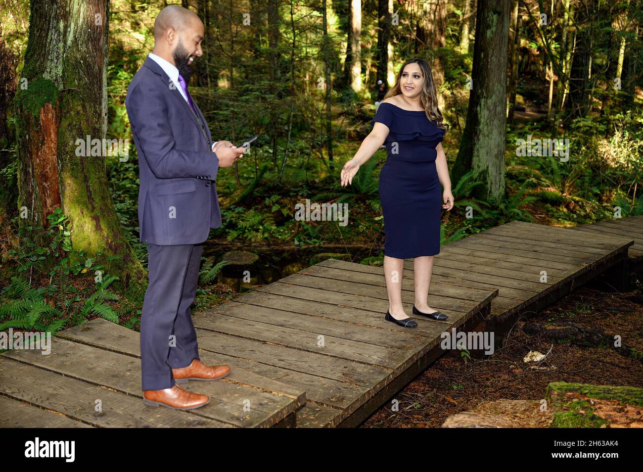 Ein junger Mann in einem Anzug und seine Verlobte, eine gut gekleidete Frau, die im Wald läuft, während der Mann anhält und etwas auf seinem Handy überprüft. Sie fragt h Stockfoto