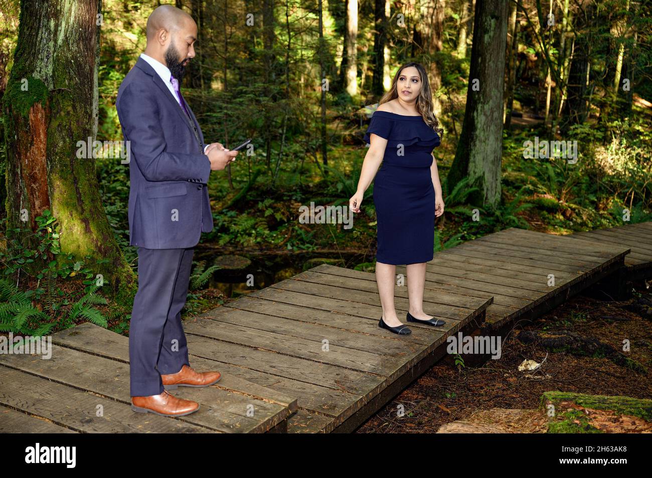 Ein junger Mann in einem Anzug und seine Verlobte, eine gut gekleidete Frau, die im Wald läuft, während der Mann anhält und etwas auf seinem Handy überprüft. Sie fragt h Stockfoto