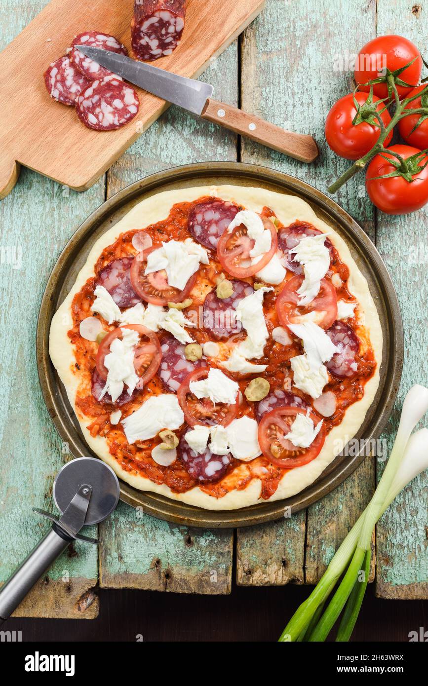 Rohe rustikale Pizza bereit zum Backen auf alten grünen Brettern. Zutaten: Tomaten, Salami, Lauch und Mozarella-Käse Blick von oben Stockfoto