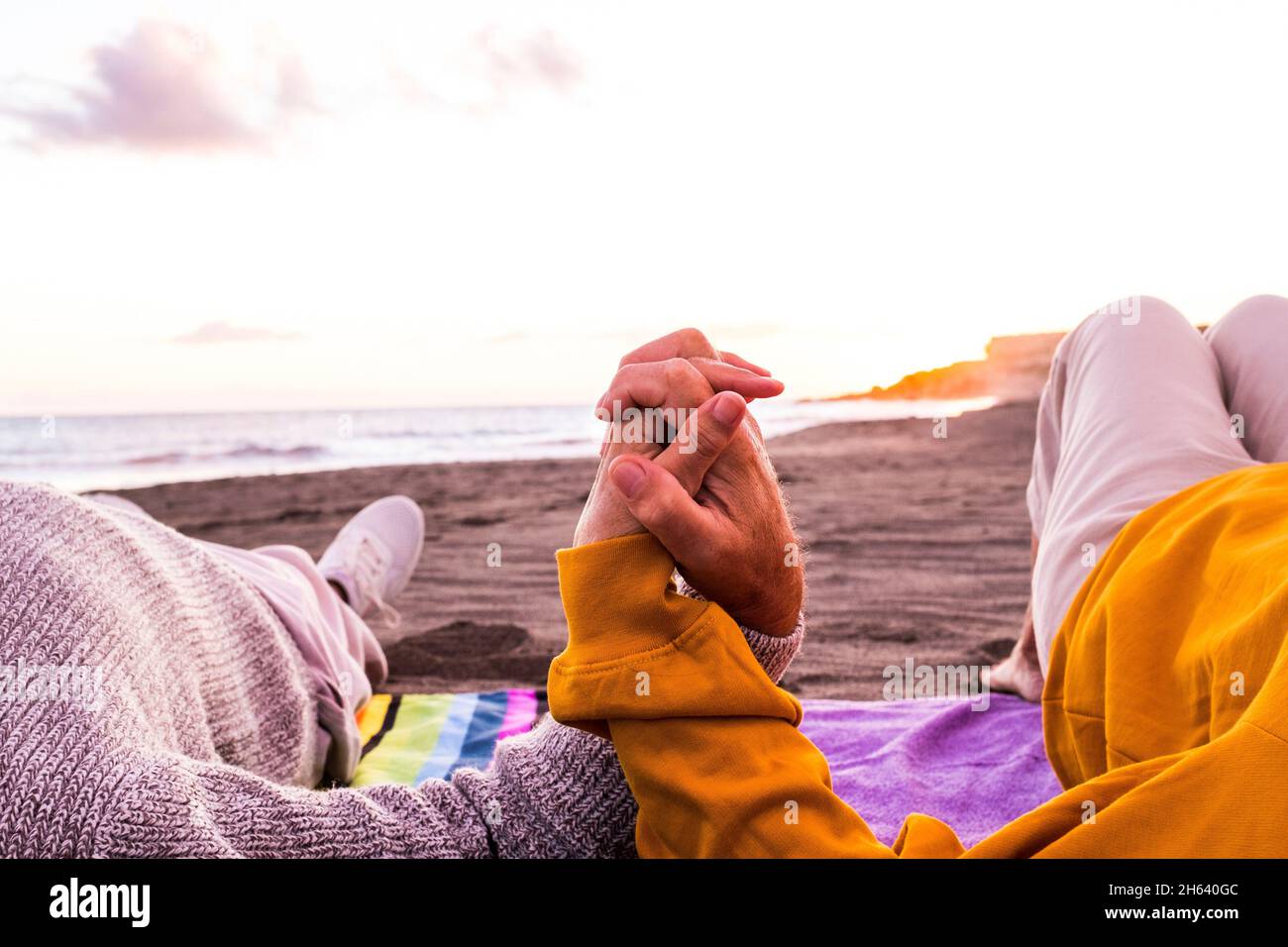 Nahaufnahme von zwei Händen, die am Strand zusammenhalten, mit dem Sonnenuntergang im Hintergrund, den Sommer genießen und Spaß zusammen haben. Ein paar Leute auf dem Sand Stockfoto