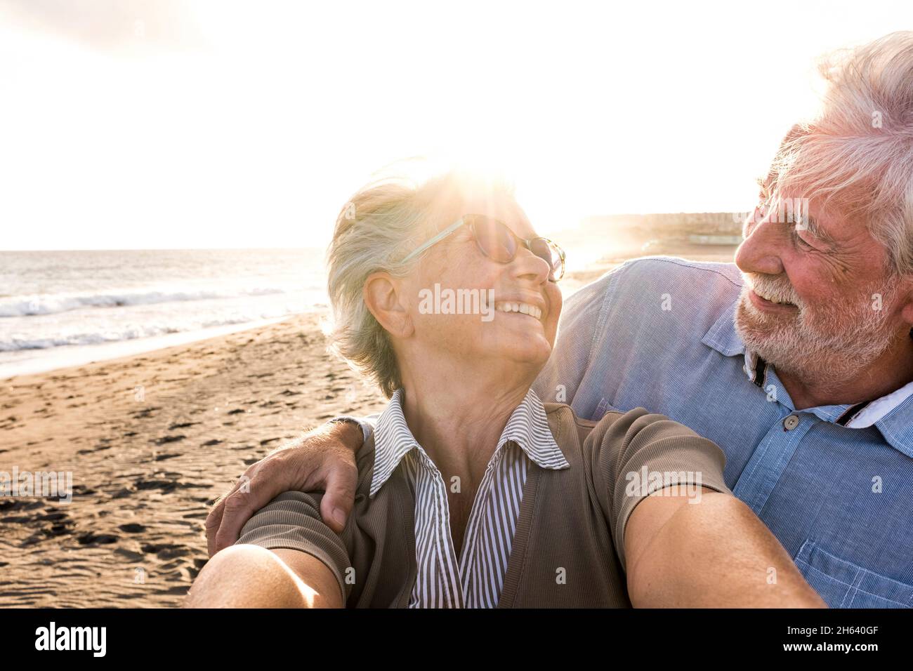 Portrait von ein paar älteren und älteren Menschen, die den Sommer am Strand genießen und auf die Kamera schauen, um ein Selfie zusammen mit dem Sonnenuntergang im Hintergrund zu machen. Zwei aktive Senioren, die im Freien unterwegs sind. Stockfoto