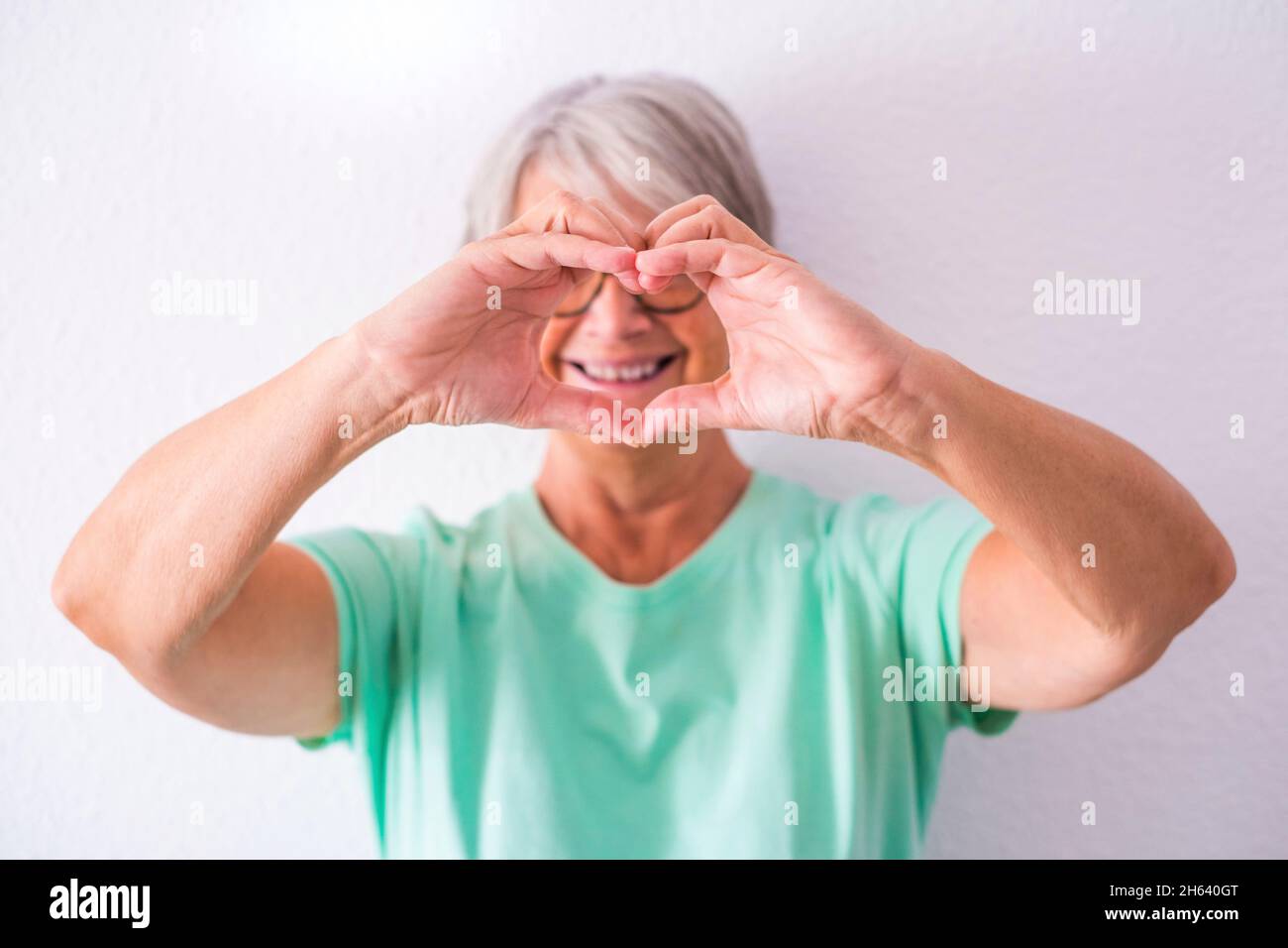 Porträt einer reifen und alten Frau, die ihnen ein Herz mit ihren Händen lächelnd und Spaß zu Hause gemacht Kamera zeigt. Weibliche Senior kümmert sich um und liebevolle Menschen Stockfoto
