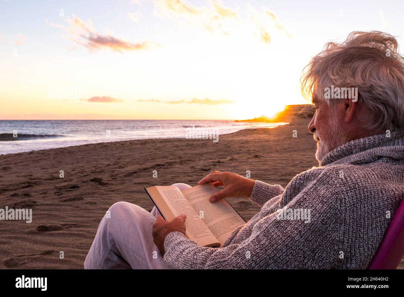 Ein reifer und alter Mann liest ein Buch, sitzt auf einem Stuhl am Strand auf dem Sand mit dem Sonnenuntergang im Hintergrund. Männliche Person genießt das Meer oder das Meer. Stockfoto