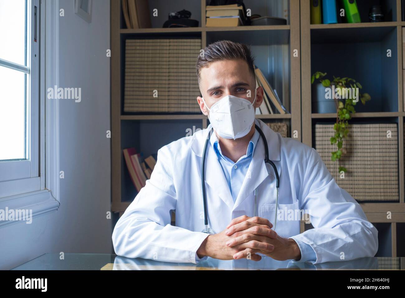 Porträt eines selbstbewussten professionellen männlichen Arztes, der eine medizinische Gesichtsmaske und eine Uniform mit Stethoskop trägt, das im Krankenhausbüro mit gekreuzten Armen steht und die Kamera betrachtet, sowie eines Gesundheitskonzepts Stockfoto