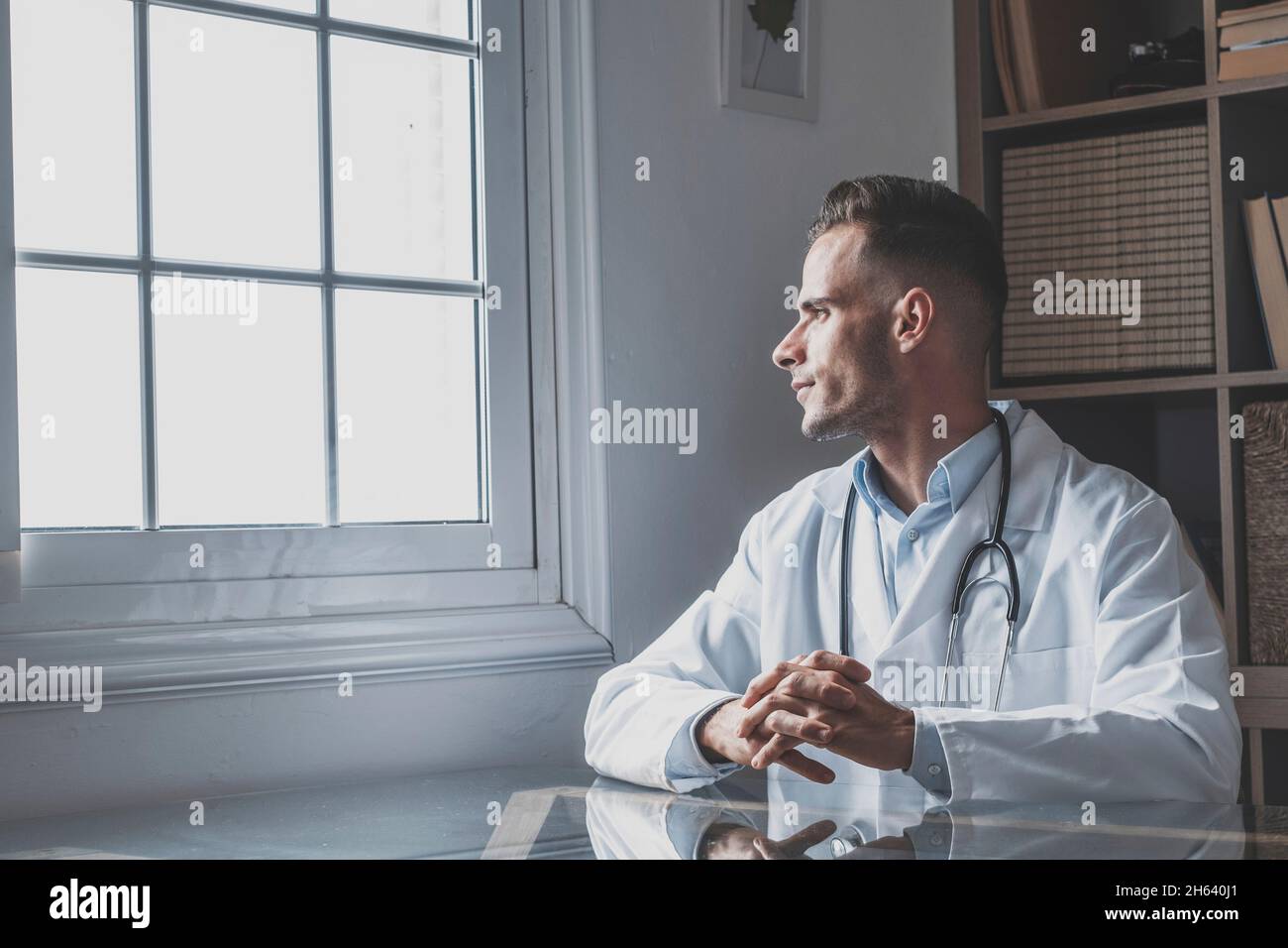 Nachdenklicher junger männlicher kaukasischer Arzt in weißer medizinischer Uniform Blick im Fenster Abstand denken oder grübeln, ernster Mann gp planen zukünftige Karriere oder Erfolg in der Medizin, visualisieren am Arbeitsplatz schriftlich Stockfoto