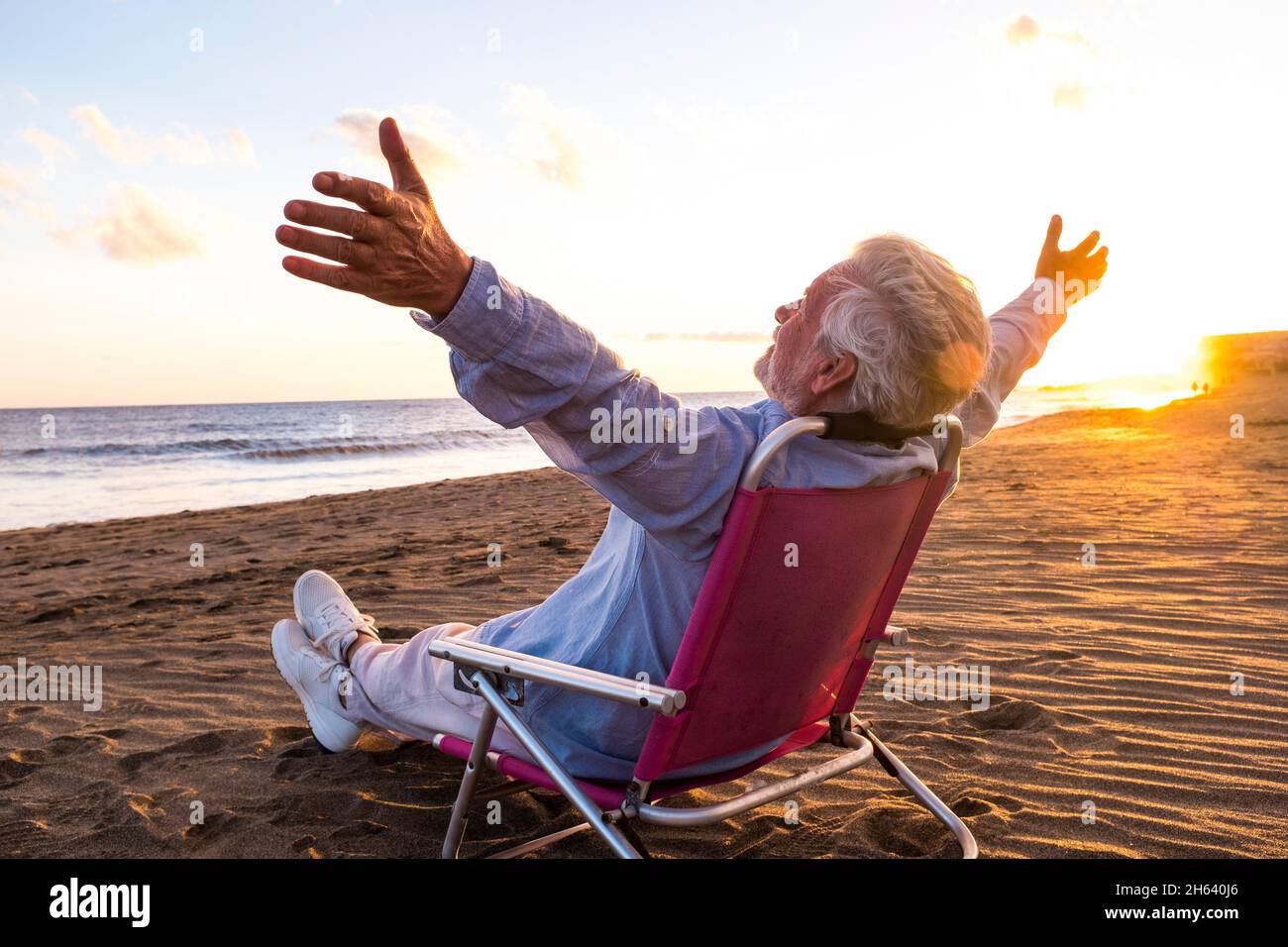 Ein reifer und alter Mann genießt die Sommerferien allein am Strand, sitzt in einem kleinen Stuhl mit Blick auf das Meer. Männliche Person fühlt sich frei mit geöffneten Armen. Freiheitskonzept. Stockfoto