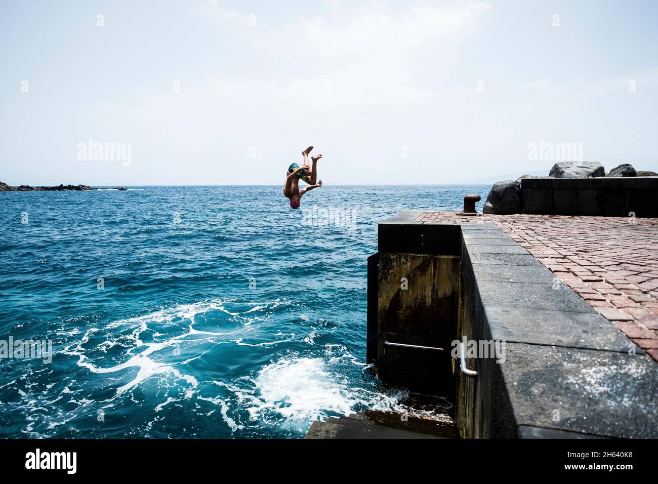 Ein junger und glücklicher Teenager, der an einem heißen Sommertag von einer Klippe zum Wasser des Meeres oder des Ozeans springt und am Strand genießt. Aktiver und schöner Lebensstil im Freien Stockfoto