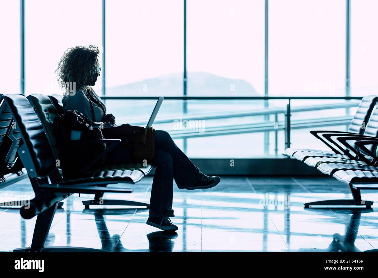 Eine Frau setzt sich allein am Flughafen-Gate nieder und wartet auf den Flug, um zu reisen - Geschäfts- oder Urlaubsreisende - Fenster mit heller Aussicht im Hintergrund - Dame, die sitzt und wartet, verzögern das Abflug-Flugzeug Stockfoto