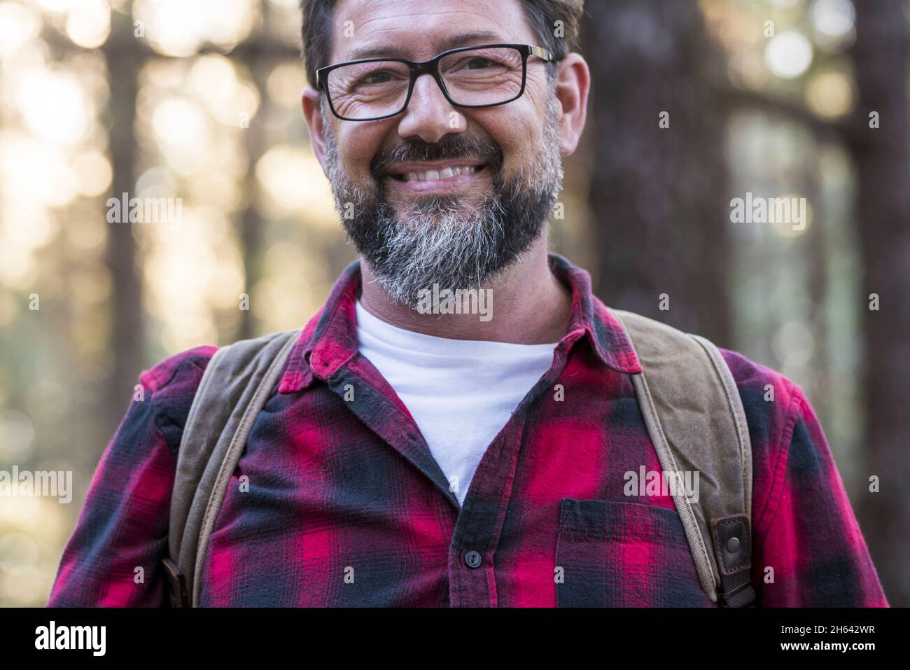 Glücklicher, gutaussehender Mann in Brillen und kariertem Hemd, der die Kamera anschaut, während er auf einer Wanderung im Wald etwas Interessantes bewundert. Mann, der im Urlaub in den Wald wandert Stockfoto Glücklicher, gutaussehender Mann in Brillen und kariertem Hemd, der die Kamera anschaut, während er auf einer Wanderung im Wald etwas Interessantes bewundert. Mann, der im Urlaub in den Wald wandert Stockfoto