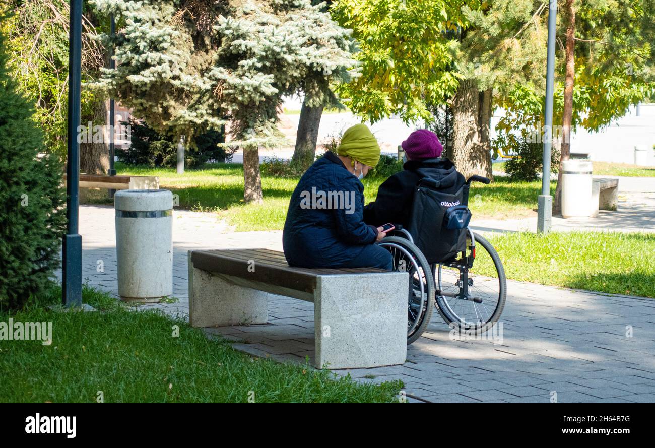 Eine ältere Frau, die sich in einem Park in Almaty um eine ältere Frau im Rollstuhl kümmert. Hilfe und Unterstützung älterer Frauen im Rollstuhl, covid-19 Lockdown Stockfoto