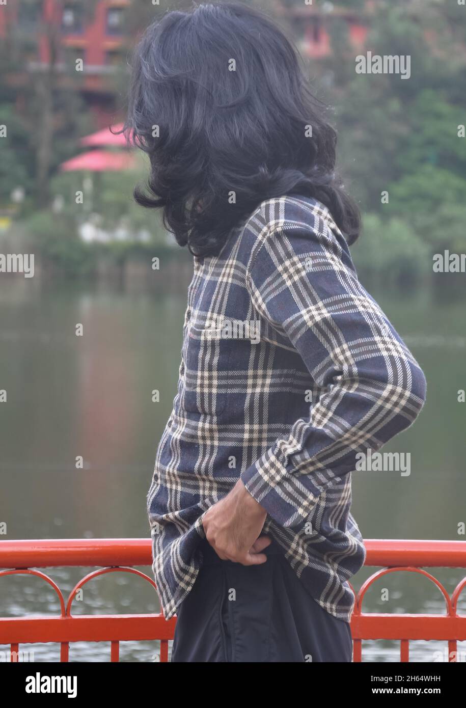 Ein junger Mann mit langen Haaren, der seine Hose justiert, während er mit einem Blick auf den See steht Stockfoto