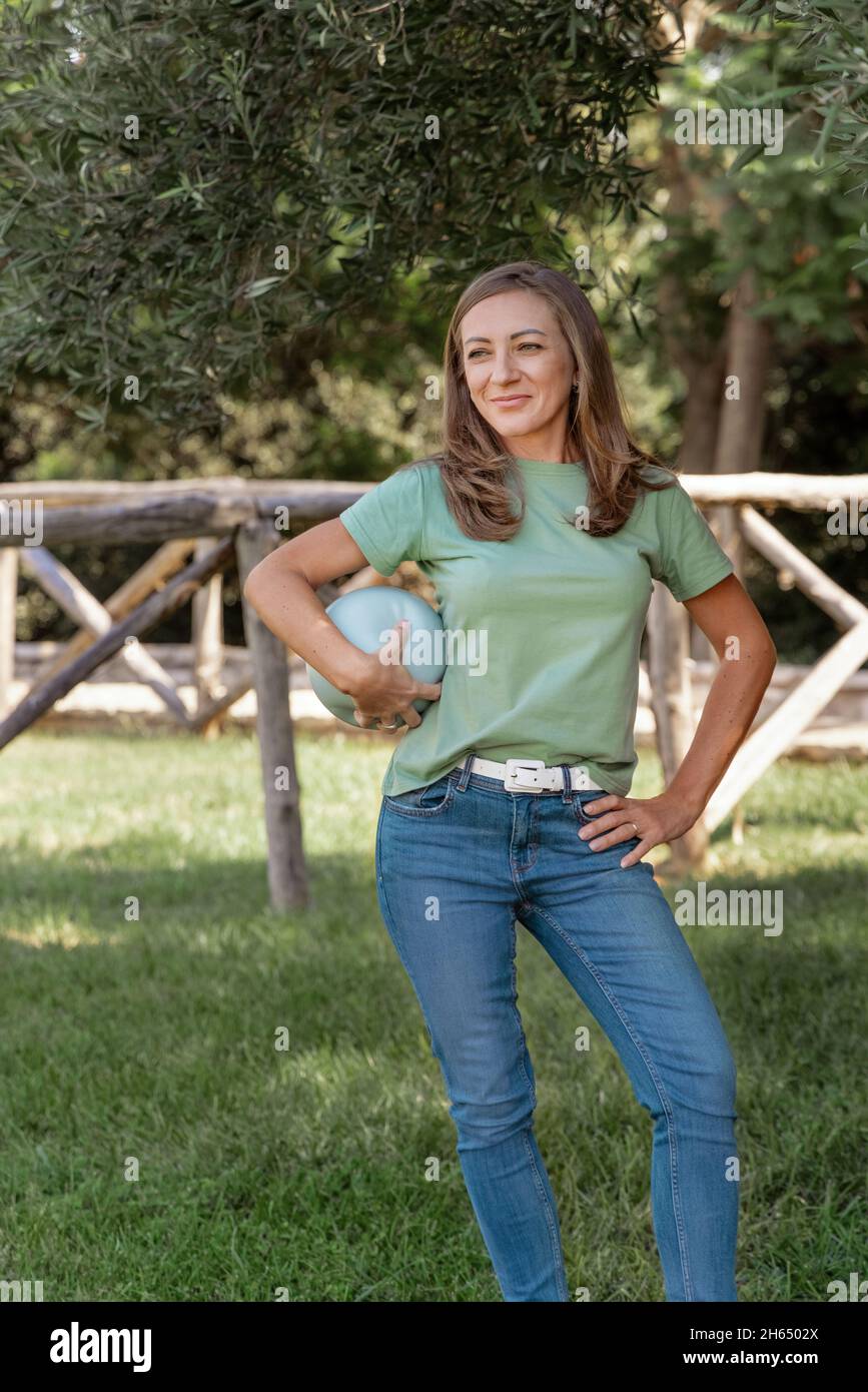 Kaukasische Frauen mittleren Alters, die T-Shirt und Jeans tragen, bleiben mit einem Ball im Park, drei Viertel Schuss Stockfoto