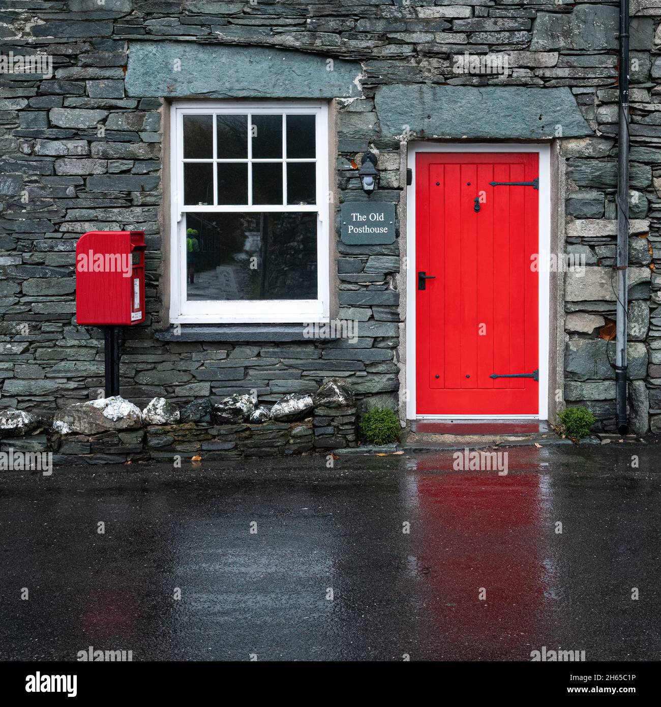 Das Old Posthouse im hübschen Lake District-Dorf Rosthwaite, Cumbria, England, Großbritannien, mit einer roten Tür, die sich auf der nassen Straße widerspiegelt Stockfoto