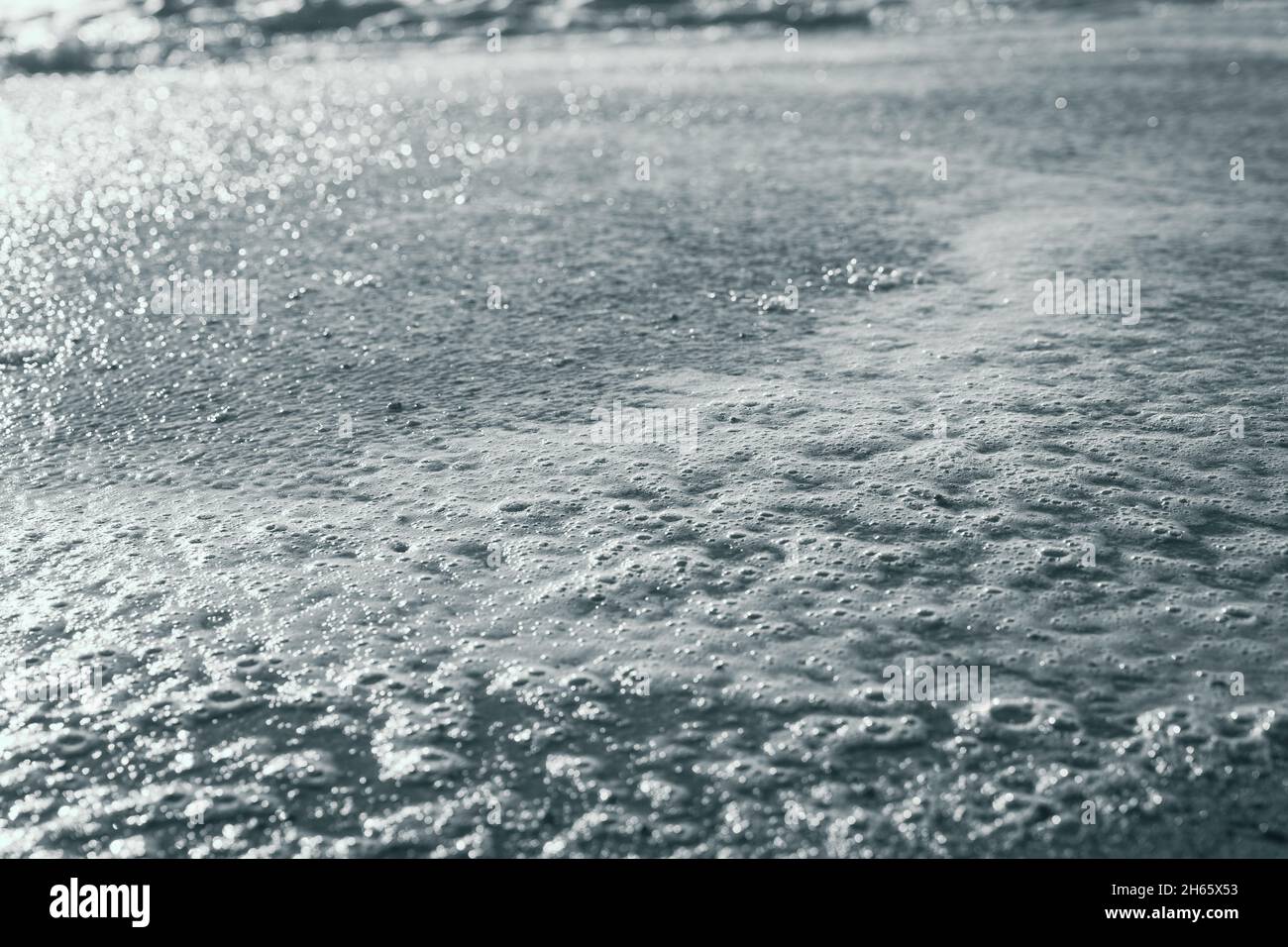 Nahaufnahme von Schaum am Meeresufer mit Sonnenreflexion auf dem Wasser. Abstraktes Hintergrundkonzept. Schwarzweiß-Fotografie Stockfoto