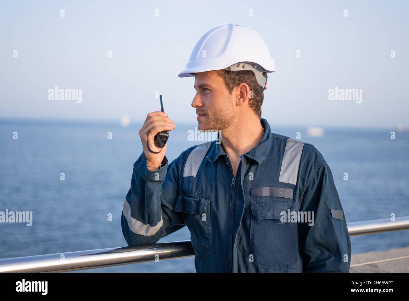 Professioneller männlicher Hafenarbeiter in Schutzuniform und Helm, der über UKW-Funkgerät kommuniziert, während er auf einem eingezäunten Damm in der Nähe des Meeres steht Stockfoto