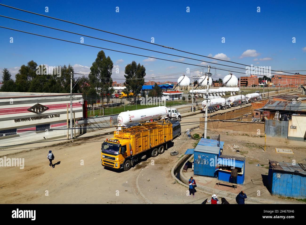 Senkata, El Alto, Bolivien. November 2021. Tankwagen für Flüssiggas und ein Lieferwagen für Inlandsflaschen (gelb) vor der Senkata-Brennstofffabrik auf der Av 6 de Marzo / Camino Oruro in El Alto. Yacimientos Petrolíferos Fiscales Bolivianos (YPFB, Boliviens staatliches Öl- und Gasunternehmen) verfügt hier über eine große Raffinerie- und Speicheranlage, Die La Paz, El Alto und die Umgebung mit Benzin, Diesel und flüssigem Erdgas (in Flaschen für den Hausgebrauch, aber auch für Fahrzeuge und andere Industrien) versorgt. Im Hintergrund sind einige der sphärischen Gasspeichertanks zu sehen. Stockfoto