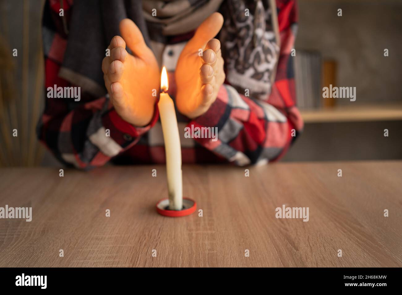 Nahaufnahme der männlichen Hände, die sich in der Nähe einer brennenden Kerze aufwärmen, die auf dem Tisch steht. Konzept der Probleme mit der Heizung, Kälte zu Hause im Winter. Stockfoto