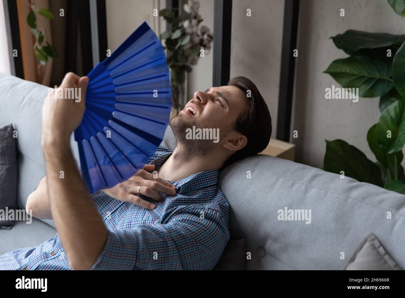 Erschöpfter junger Mann, der zu Hause einen Papierventilator benutzt. Stockfoto