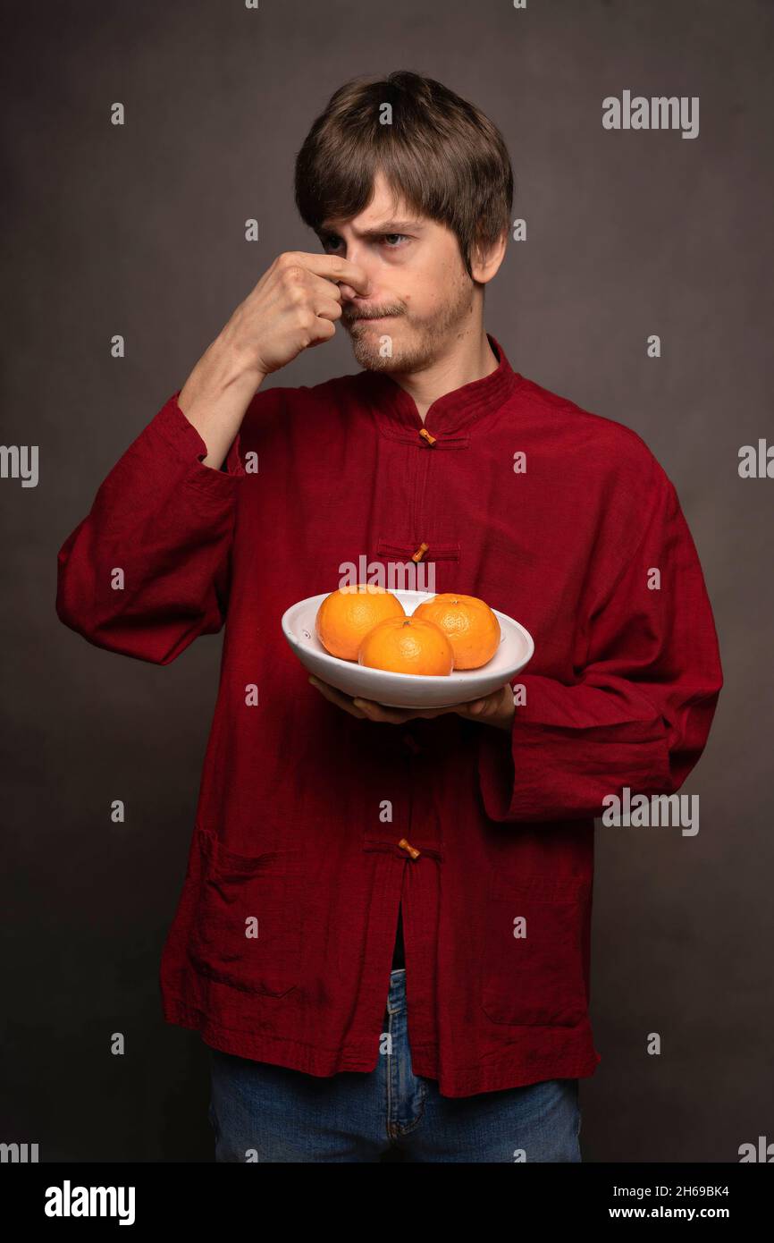 Junger, hübscher, großer, schlanker weißer Mann mit braunem Haar, das reeking Orangen in einem roten Hemd auf grauem Hintergrund hält Stockfoto