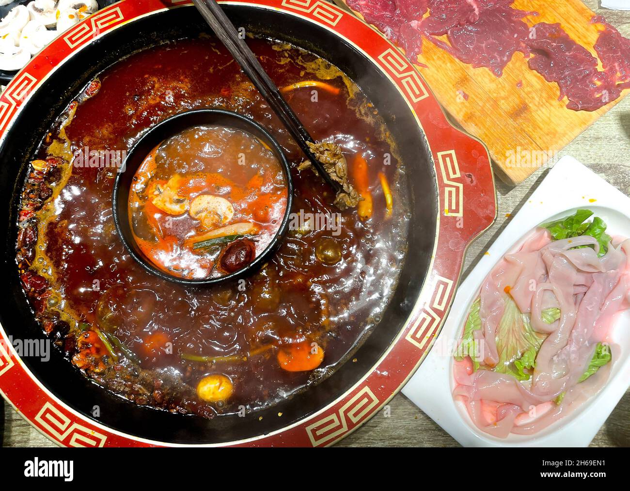 Sichuan Hotpot, Essstäbchen, die ein Stück Rindfleisch über zwei gewürzte, würzige und Tomatentopf halten, mit Gerichten von Rind- und Entendarm Stockfoto