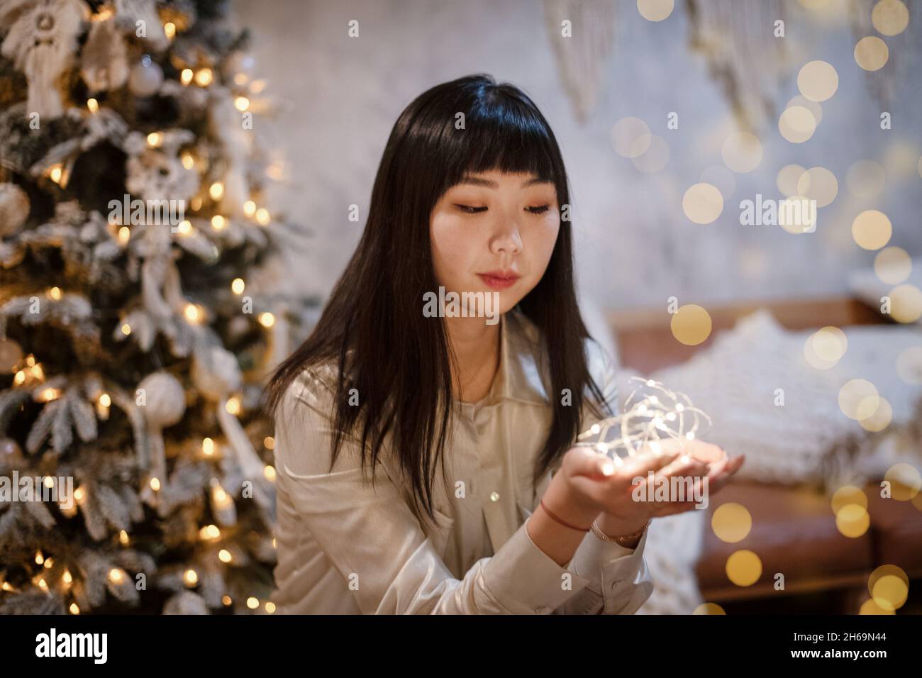 Eine junge, schöne koreanisch-asiatische Frau hält eine beleuchtete festliche Girlande in ihrem gemütlichen Zuhause mit Träumen von positiven Emotionen in der Nähe des Weihnachtsbaums in der Stockfoto