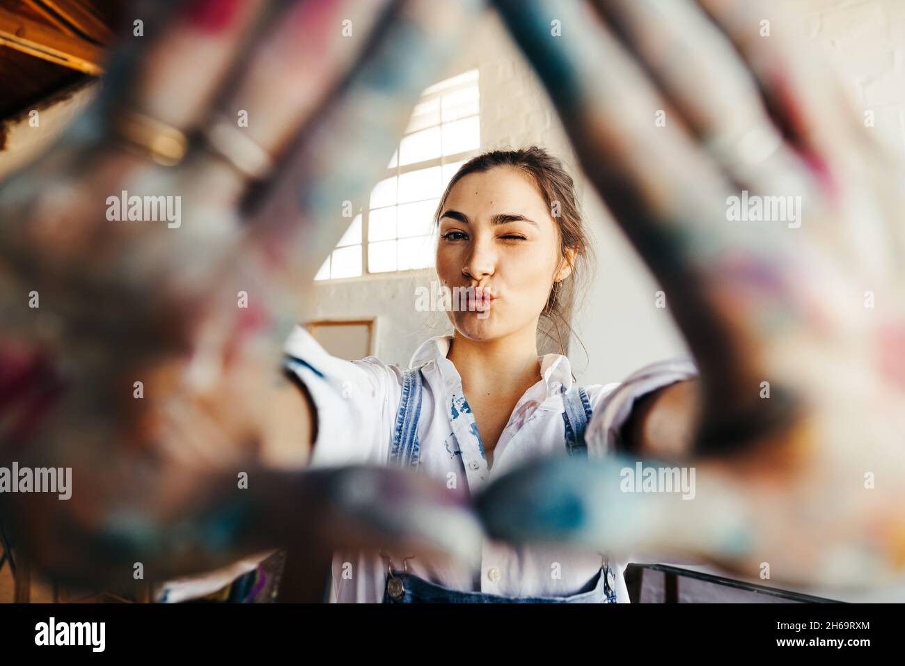 Eine Künstlerin umrahmt ihr Gesicht mit ihren Händen. Unbeschwerte Malerin winkend an der Kamera, während sie in ihrem Kunststudio steht. Kreative junge Frau ma Stockfoto