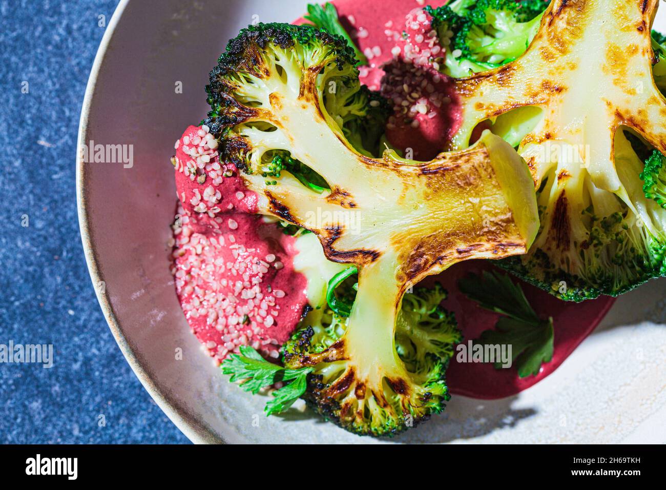 Gegrilltes Brokkoli-Steak mit rosa Rote-Bete-Sauce und Hanfsamen in weißer Schüssel, blauer Hintergrund, Nahaufnahme. Gesundes veganes Lebensmittelkonzept. Stockfoto