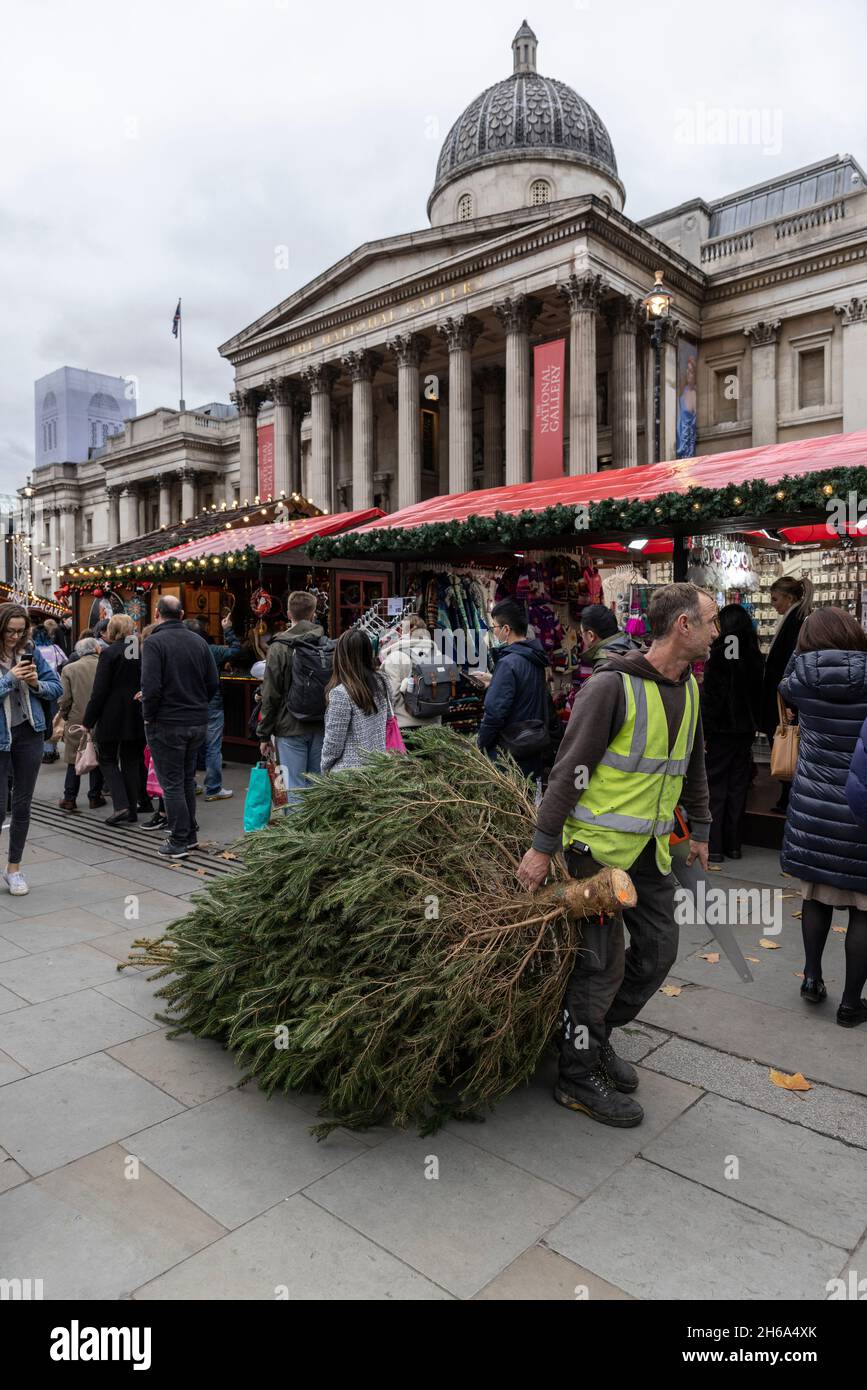 Ein Arbeiter trägt einen Weihnachtsbaum auf dem Weihnachtsmarkt, Trafalgar Square, zu Beginn der Weihnachtszeit 2021 in der Hauptstadt London, England. Stockfoto