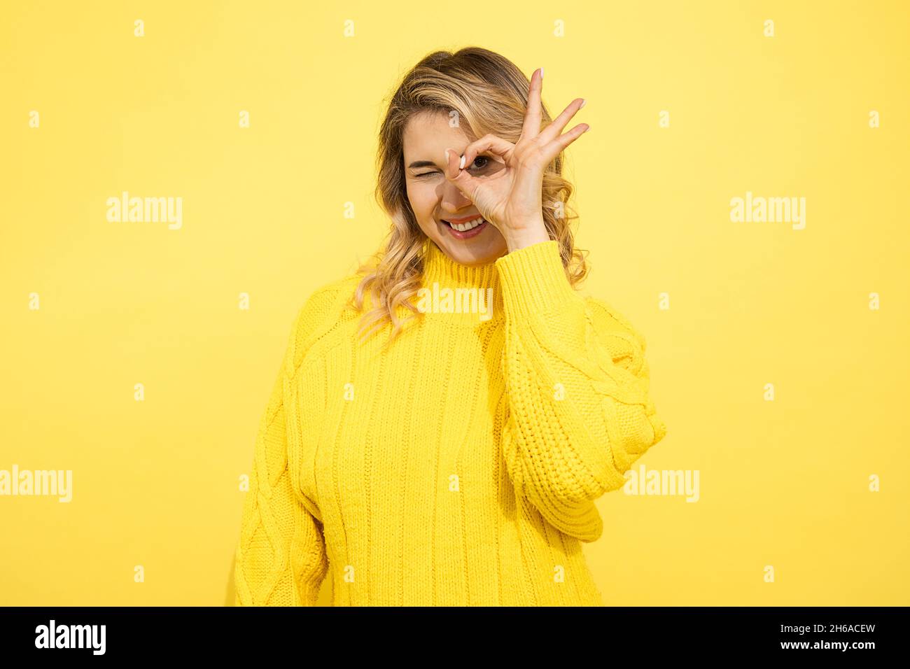 Junge schöne blonde Frau in gestricktem Pullover schaut durch ok Zeichen auf ihre Finger, Hände. Lächelnd auf Kamera, glücklich auf gelbem Hintergrund, Studio Sho Stockfoto