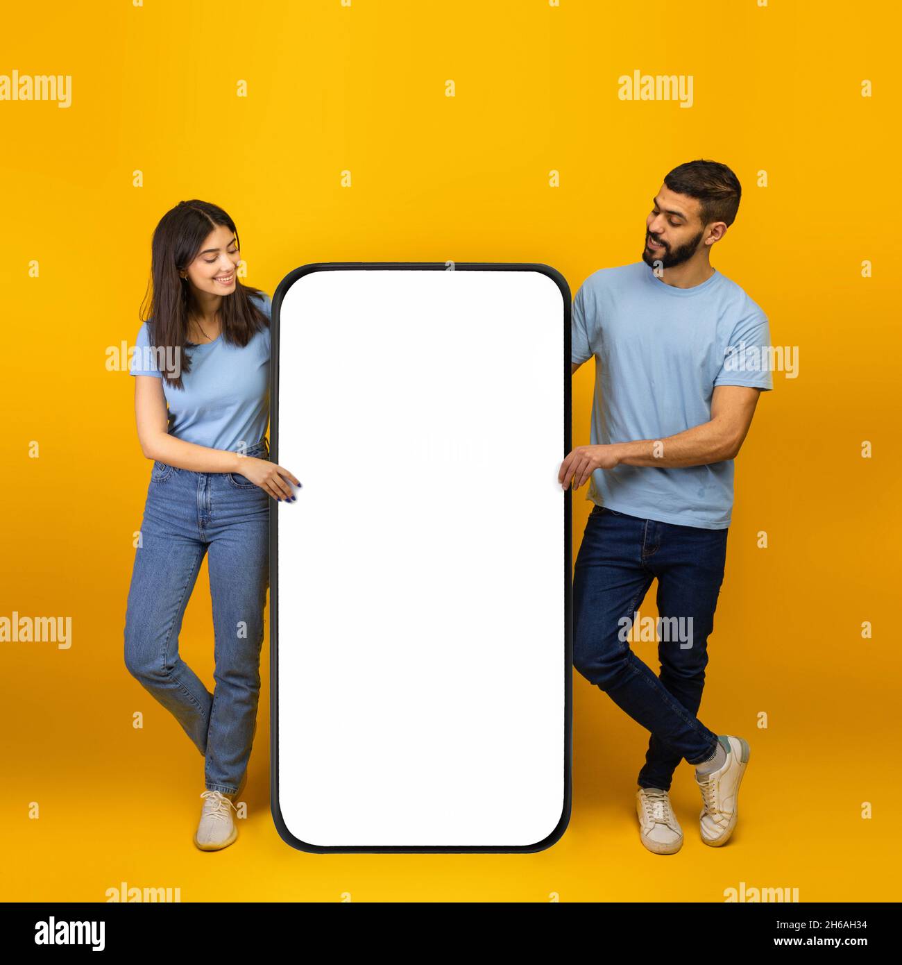Coole mobile App. Tausendjährige arabische Ehegatten stehen in der Nähe eines riesigen Smartphones mit Mockup, bewerben App oder Website, Handy mit weißem leeren Bildschirm, brüllen Stockfoto
