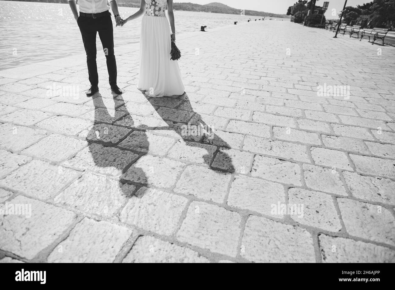 Braut und Bräutigam halten die Hände und stehen auf den Pflastersteinen am Meer. Schwarzweiß-Fotografie Stockfoto