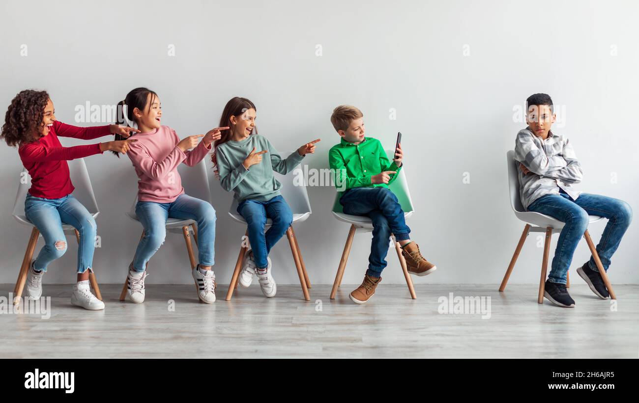Mobbing Und Cybermobbing In Der Schule. Jugendliche Kinder Lachen Über Opfer Afrikanischer Amerikanischer Junge Mit Smartphone Sitzen Über Grauer Wand Im Klassenzimmer In Stockfoto