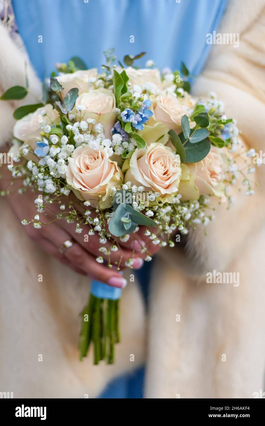 Winter Brautstrauß in den Händen. Beige Rosen, blaue und graue Blüten. Blaues Kleid und beigefarbener Pelzmantel auf der Braut. Hochzeitsbouquet in Pastellfarben aus nächster Nähe. Blumen Stockfotografie