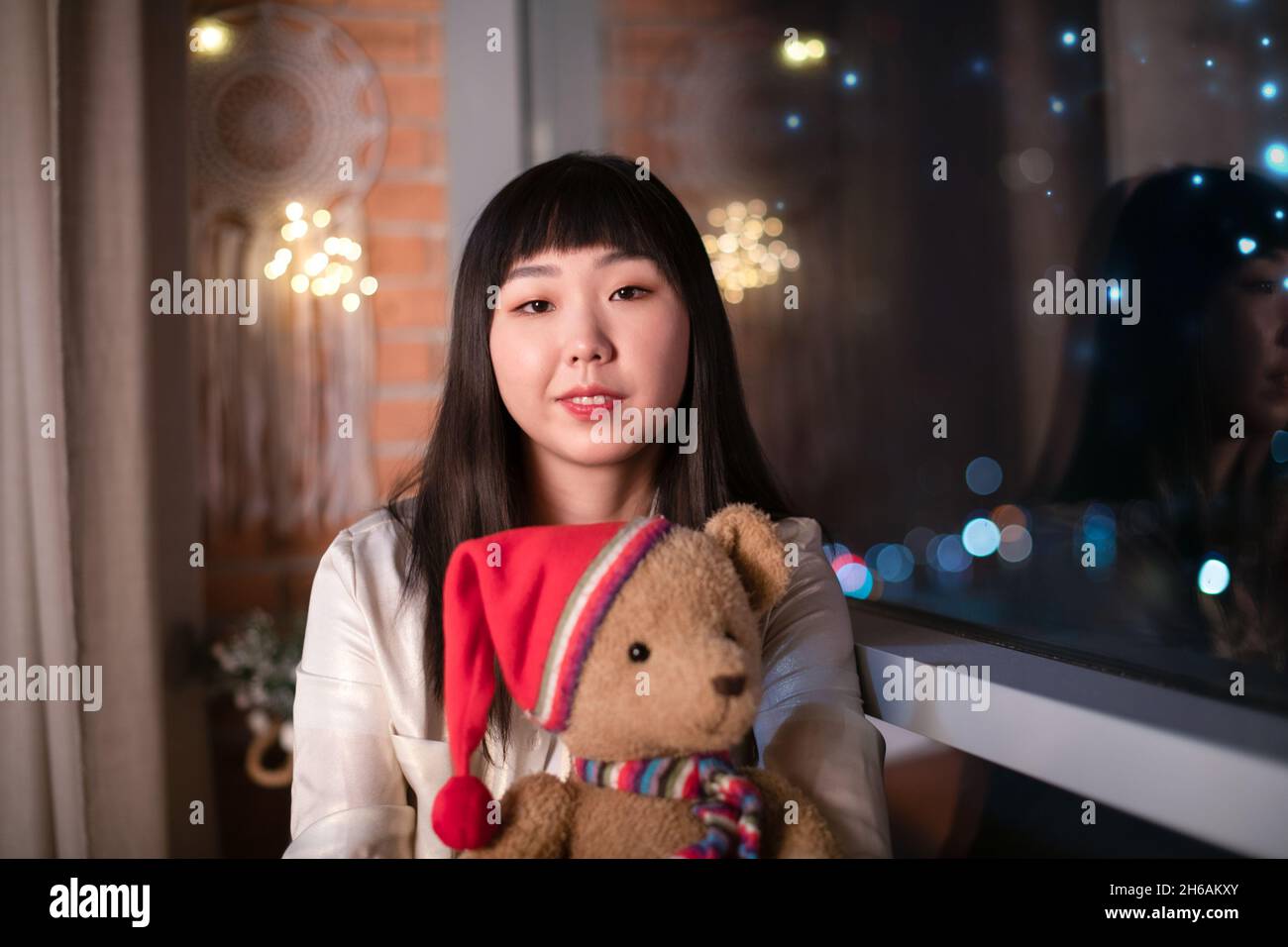 Eine niedliche einsame junge koreanische Asiatin hält einen lustigen weichen Bären in ihren Armen und sitzt auf dem Fenster am Nachtfenster des Hauses. Das Konzept des Neuen Stockfoto