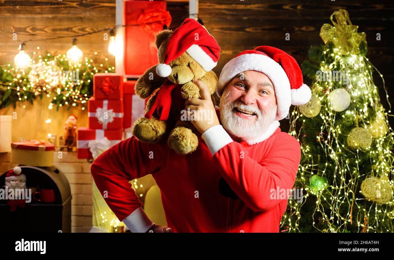 Weihnachtsmann mit Teddybär auf den Schultern. Weihnachtsferien. Bärtiger Mann in Weihnachtsmann-Kostüm in der Nähe von geschmücktem Neujahr-Baum. Stockfoto